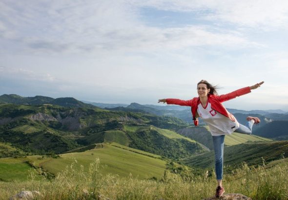 A woman joyfully balances on a rock amidst rolling green hills under a clear blue sky.